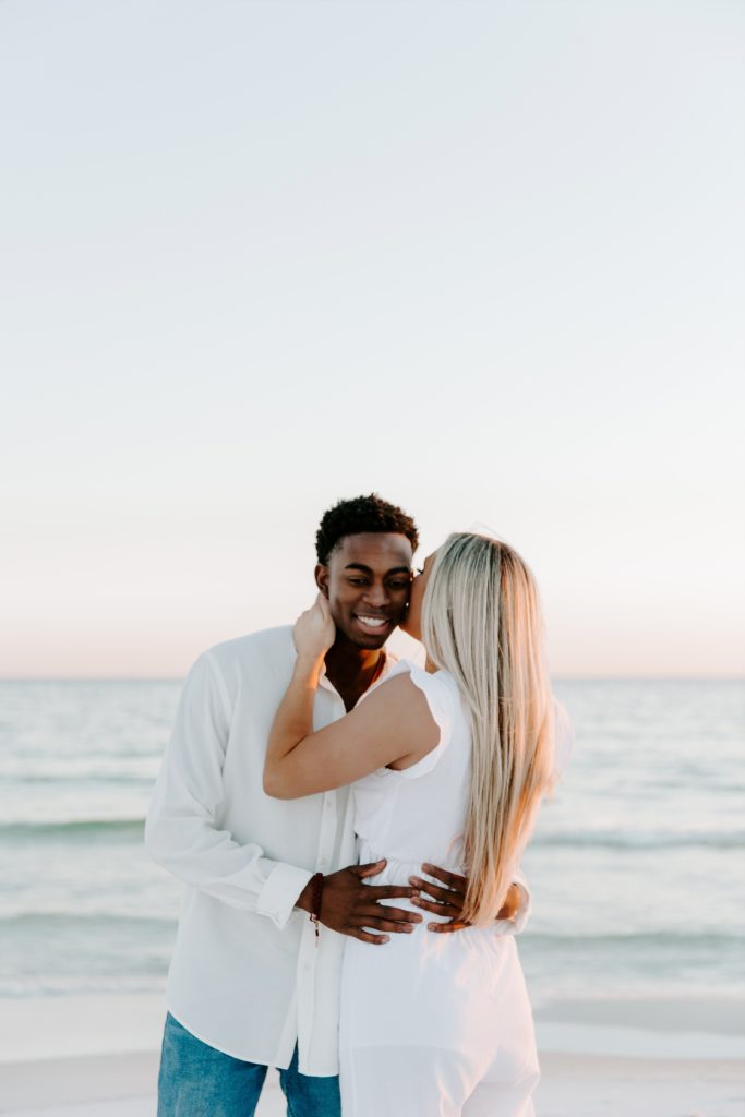 Woman giving her partner a kiss on the cheek while he has his arms wrapped around her and she has her hand on his neck during their beach couple photos in Destin, Florida