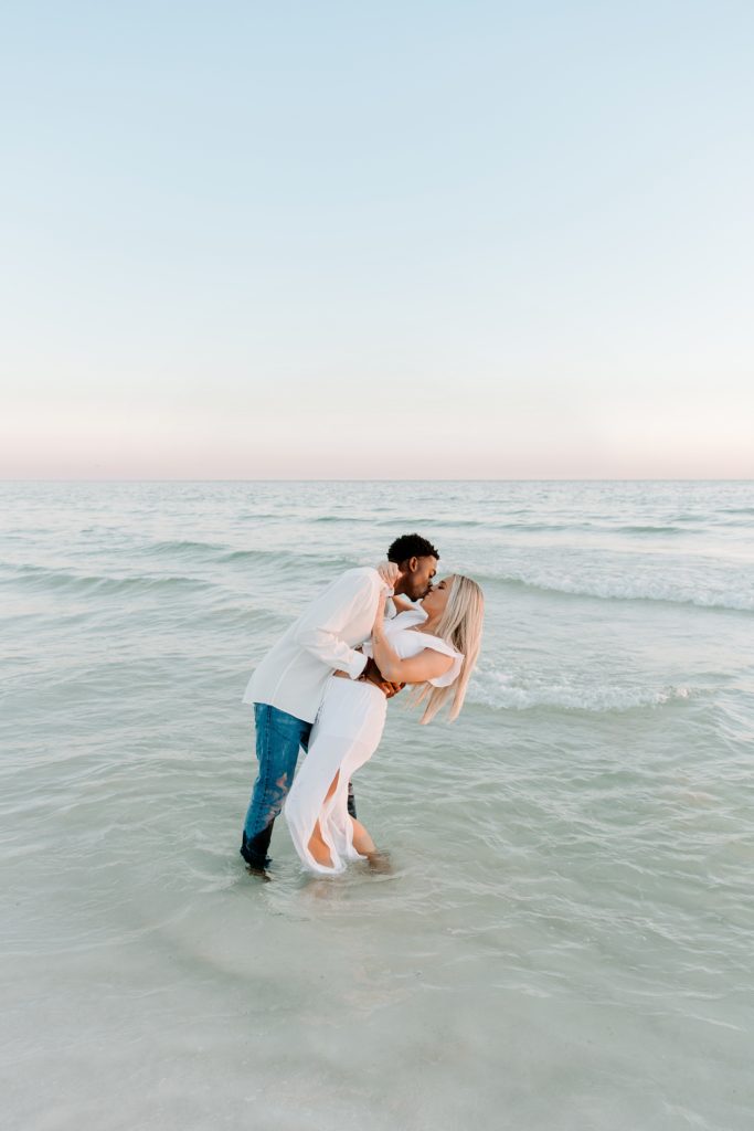 A man dipping his partner into a kiss as they are standing in the ocean at sunset during their beach couple photos in Florida