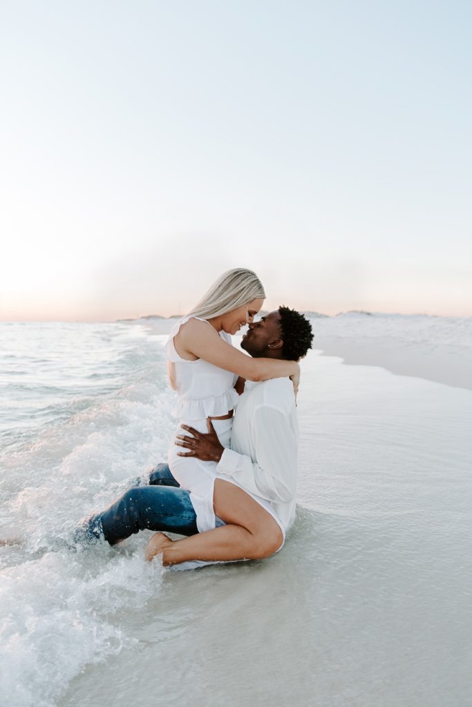 A man sitting in the ocean water with his partner sitting on top of him straddling him as they look at each other and the water is rushing around them during their beach engagement photos in Destin, FL