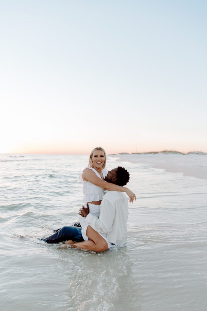 Woman sitting on her partners lap as they sit in the ocean and the woman has her arm resting on her partners shoulder and is looking off into the distance smiling during their beach couple photos in the Florida panhandle