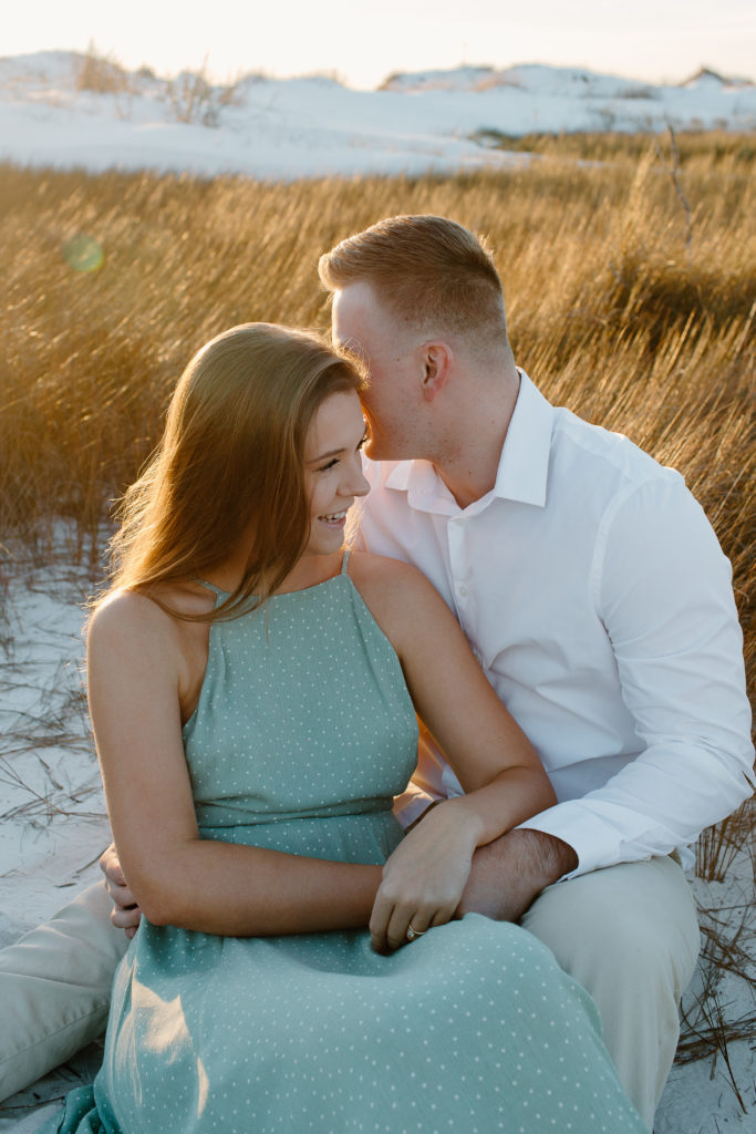 Man whispering into is partners ear as they are sitting surrounded by dune grass as the woman is smiling during their Destin, Florida engagement photos