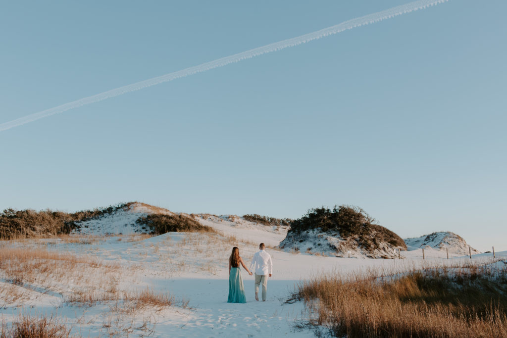 Couple walking towards large sand dunes as the sun is setting in the background during their golden hour engagement photos