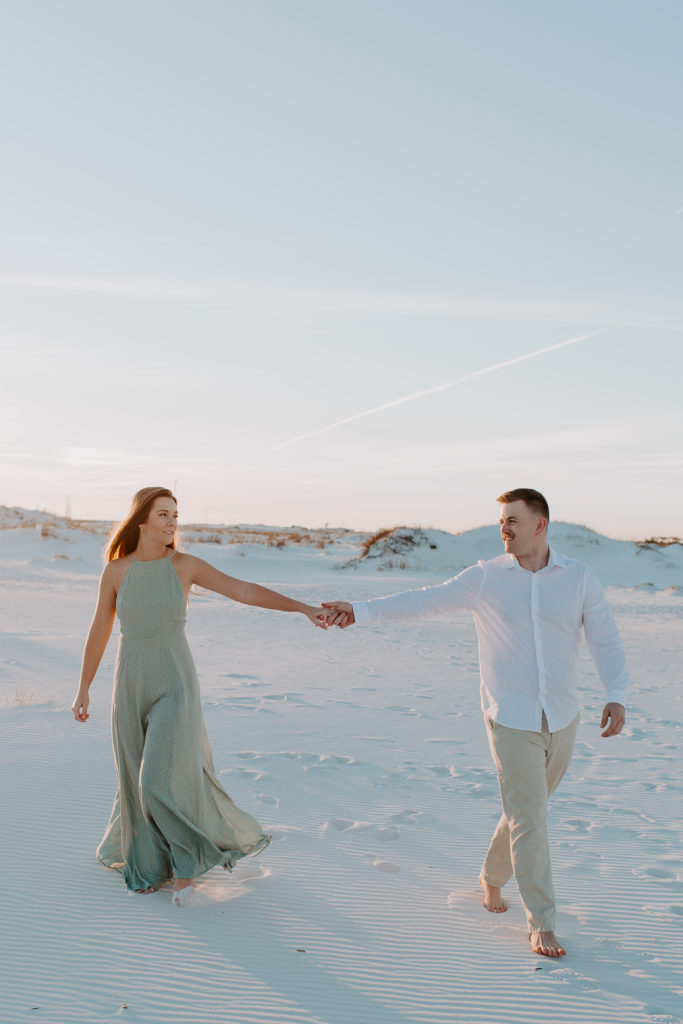 A couple holding hands and walking far apart from each other as they look at the other during their beach engagement photos