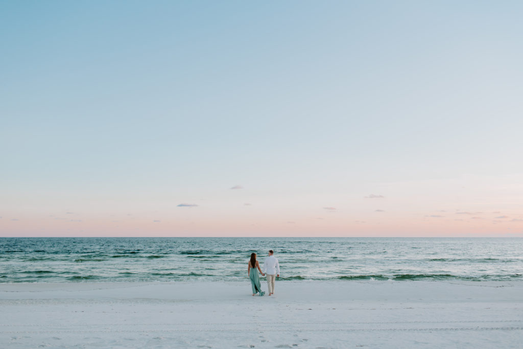 Man and woman holding hands and walking towards to the ocean as the sun is setting in the Florida panhandle