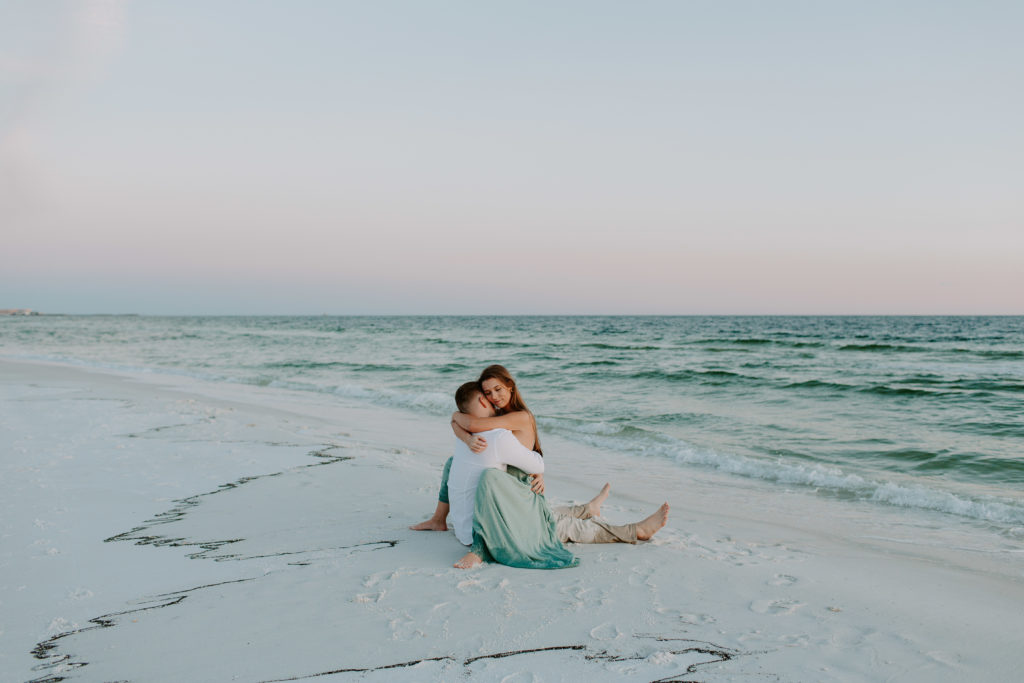 Couple sitting on the beach with the woman in the mans lap as they hug onto each other during their beach engagement photos