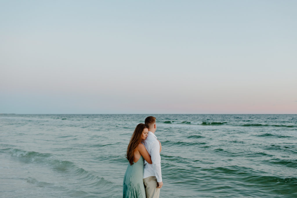 Couple standing in the water as the waves wash over them with the woman hugging the back of her partner during their golden hour engagement photos in Florida