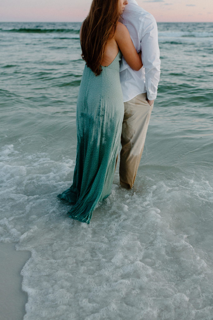 The ocean running over a couples feet as the woman holds onto her partner and rests her head on his back during their beach engagement photos in Florida