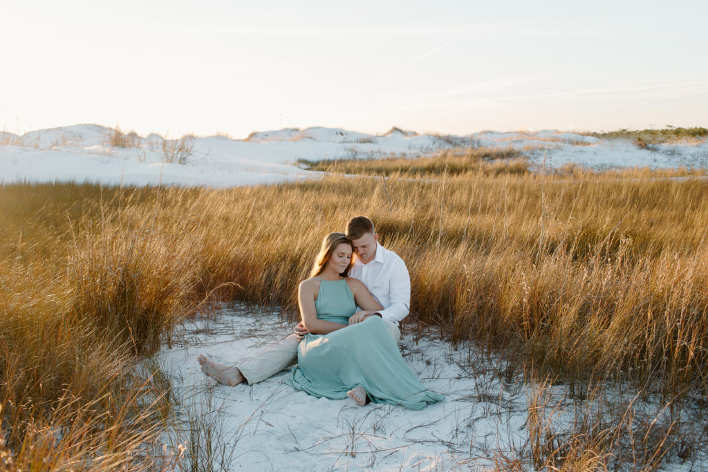 Couple sitting in the midst of dune grass with the couple leaning their heads together during their beach engagement photos in Destin, Florida