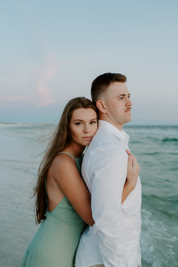 Woman hugging onto her partner as he looks off into the distance and the wind blowing through the woman's hair during their beach engagement photos in Destin