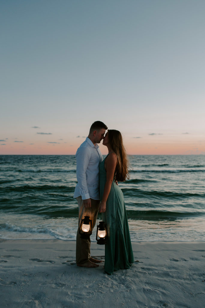 Couple standing along the waters edge with their nose touching as they are holding lanterns during their blue hour engagement session in Florida