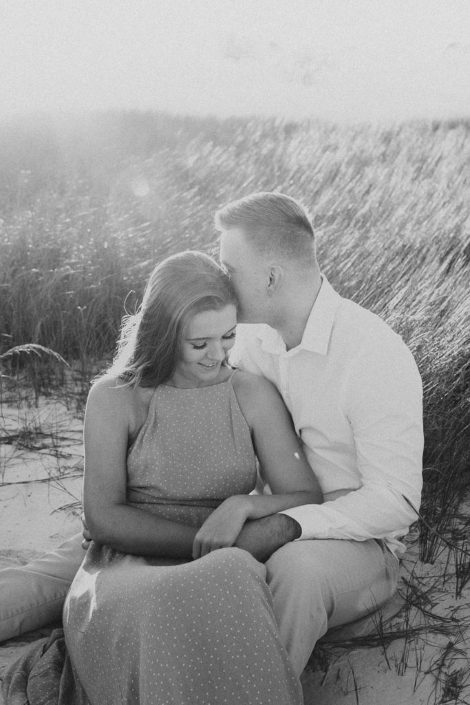 Man whispering into is partners ear as they are sitting surrounded by dune grass as the woman is smiling during their beach engagement photos