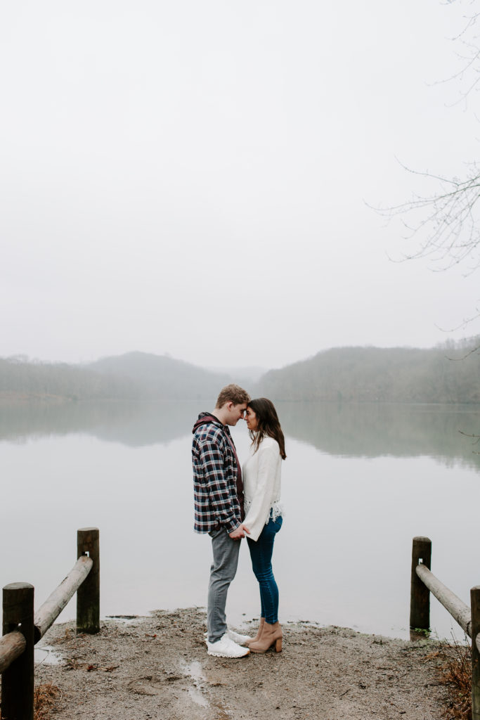 A man and a woman leaning their heads against each other while holding hands in front of a lake with fog in the background during their couple photos