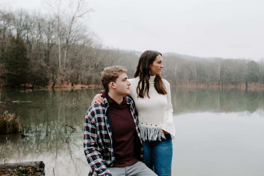 Man sitting on a fence as his partner is standing next to him with her arm around his shoulders and they are both looking off into the distance during their Nashville engagement photos