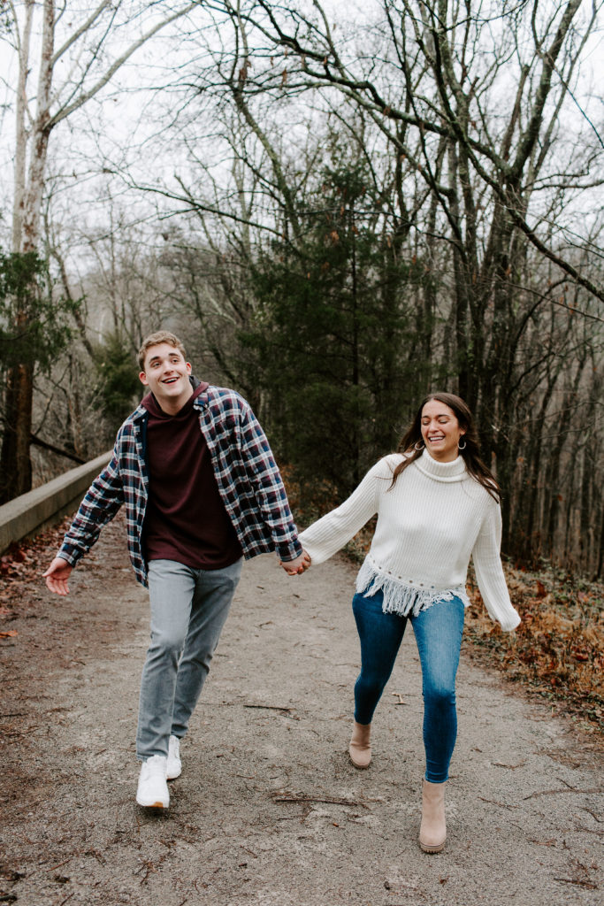 Couple holding hands and walking down a path as they are laughing during their engagement photos in Nashville, TN