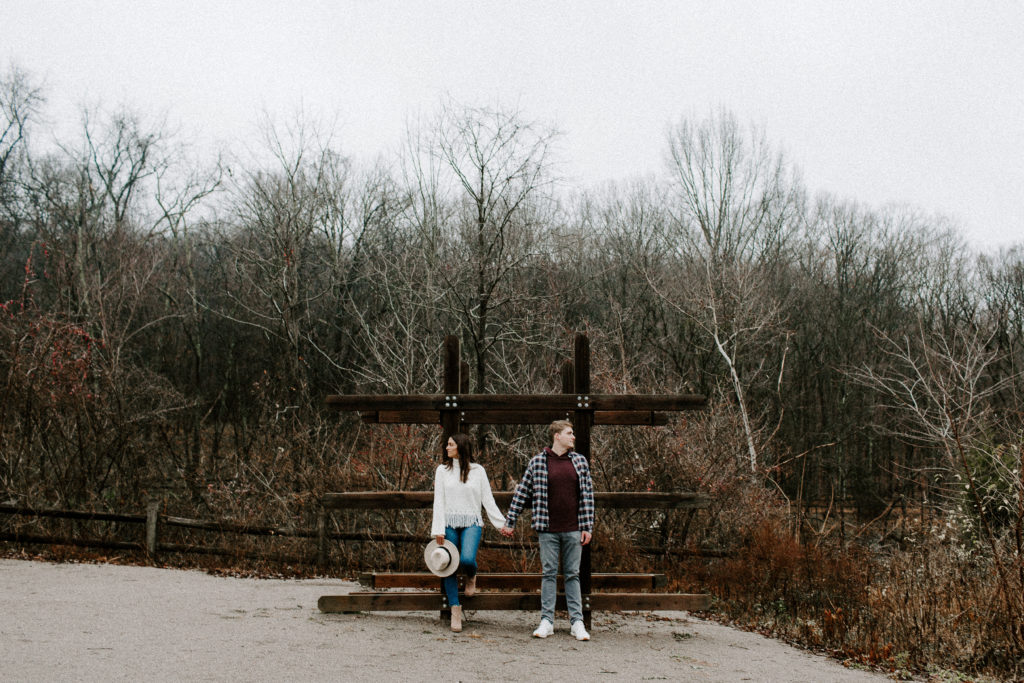 Couple standing hand in hand looking opposite directions of each other as the woman is holding a hat during their state park engagement photos in Tennessee