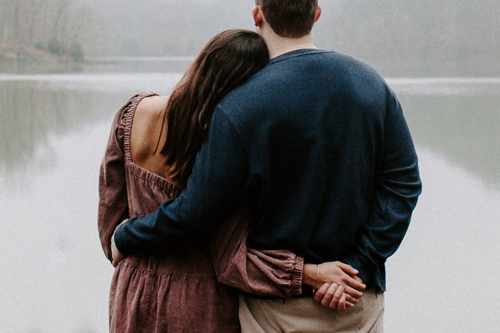 Couple looking off at the water as the woman has her arm around her partners back and they are holding hands and she is leaning her head against him during their engagement photos