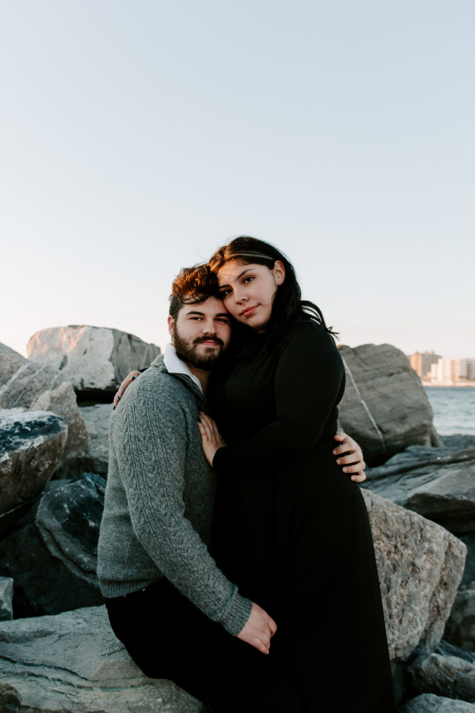 A man sitting on rocks as his partner leans into him between his legs and her hand is resting on his chest during their beach engagement photos on 30A