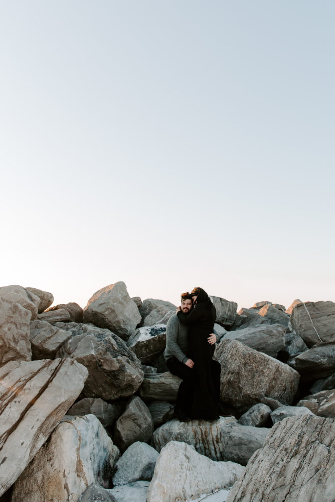 Man sitting at the base of rock jetties and his partner is standing next to him cuddling into him as the sun is setting during their Destin couple photos