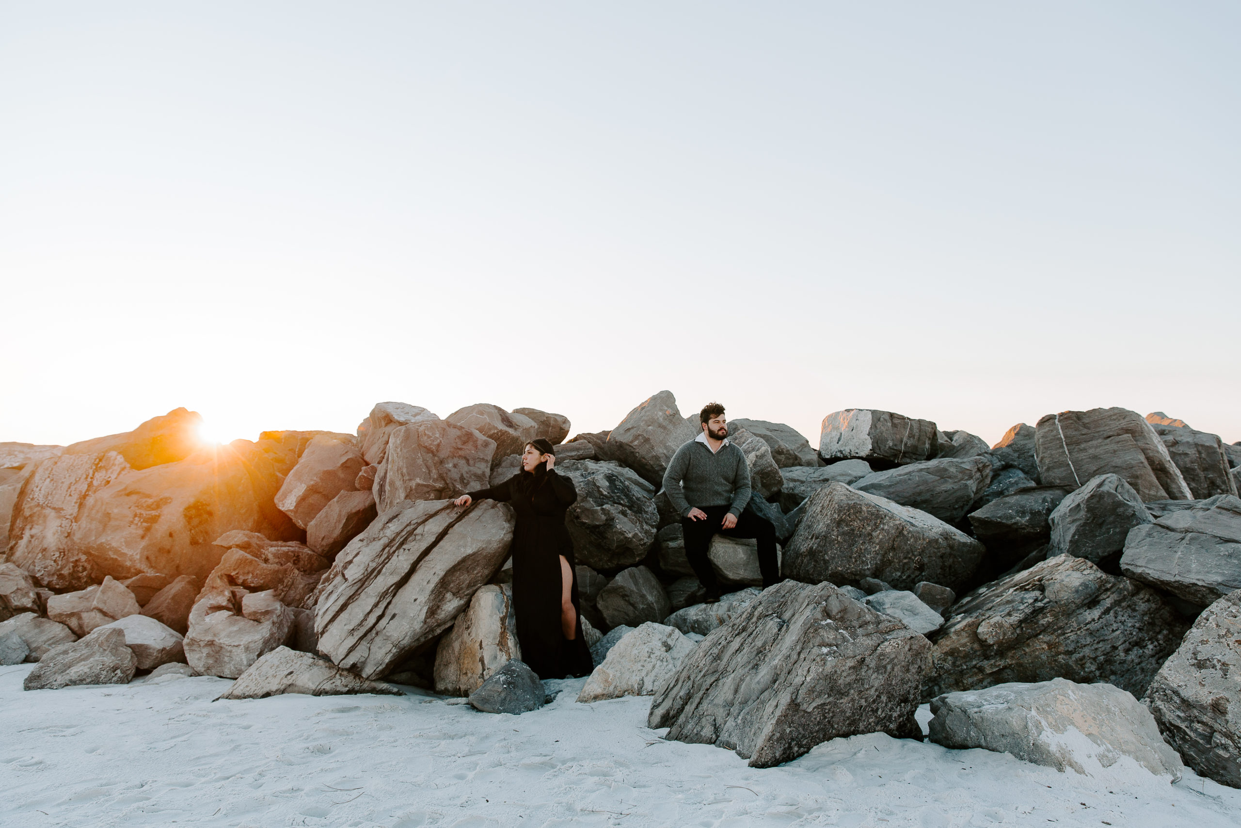 Man sitting on rocks while propping his foot on one and a woman standing a few feet away from him while brushing her hair out of her face as the sun is peaking over the rocks during their Destin couple photos in Florida