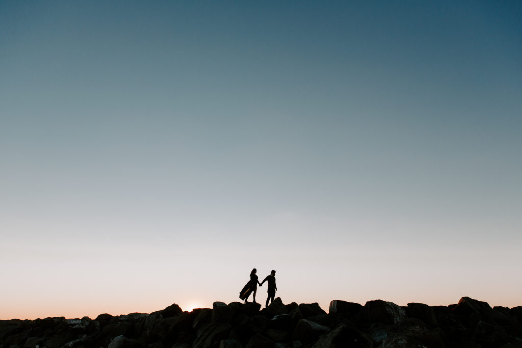 The silhouette of a couple holding hands and walking along the top of rocks as the sun is setting during their beach engagement photos in Florida