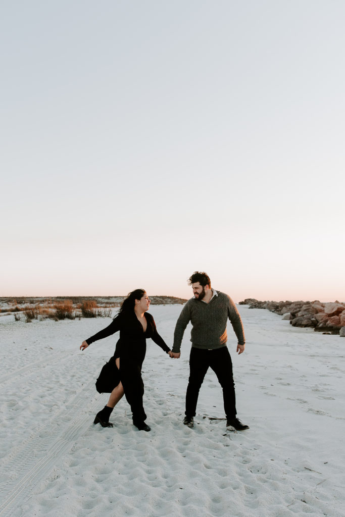 Woman and her partner holding hands and looking at each other as they are walking and the sun is setting during their beach engagement photos