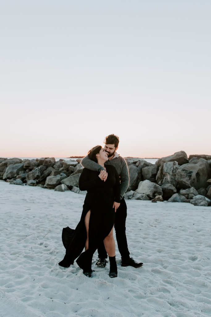 Man holding his partner from behind as she looks up at him and her dress is blowing in the wind during their golden hour couple photos in Florida