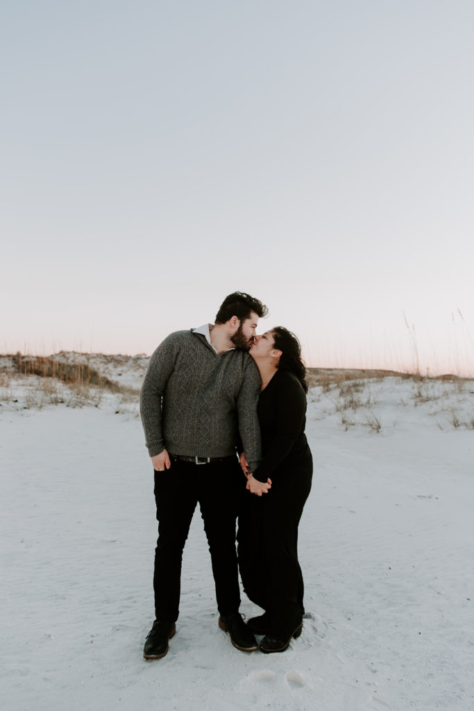 Couple standing in the midst of sand dunes as the woman is holding her partners hand and they are sharing a kiss