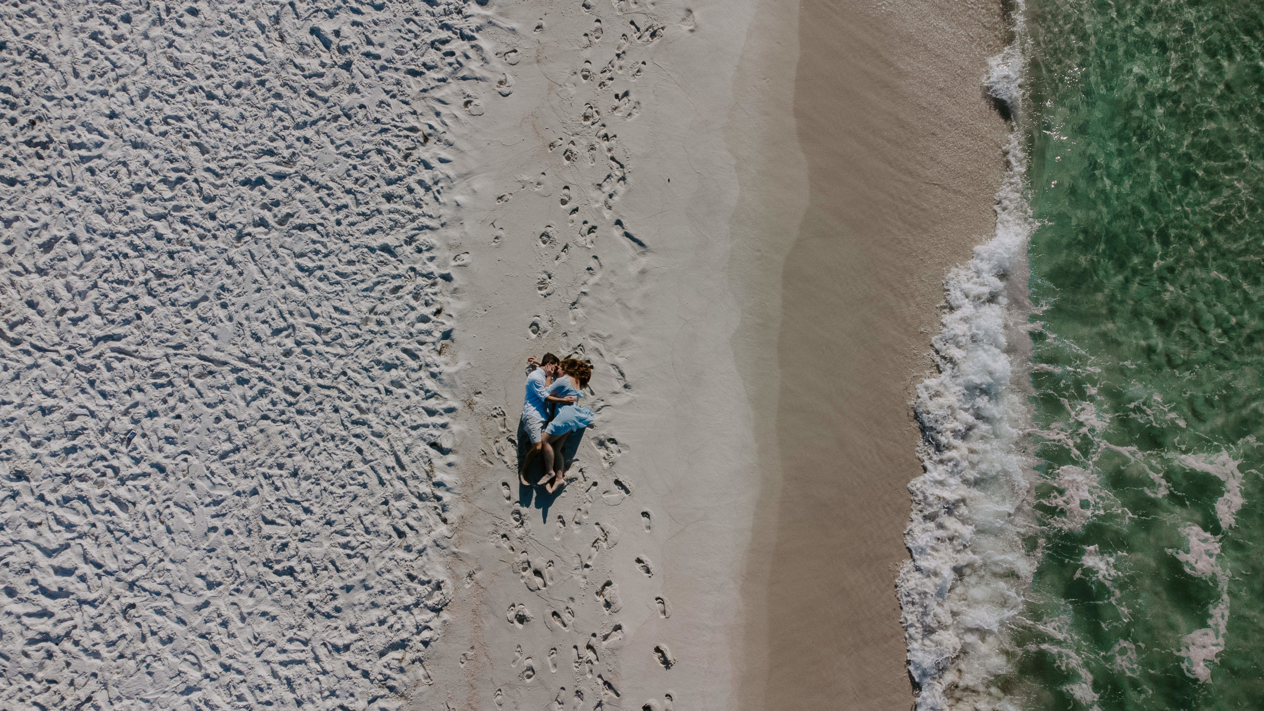 Couple laying on the beach alongside the ocean as it is coming up during their beach engagement photos