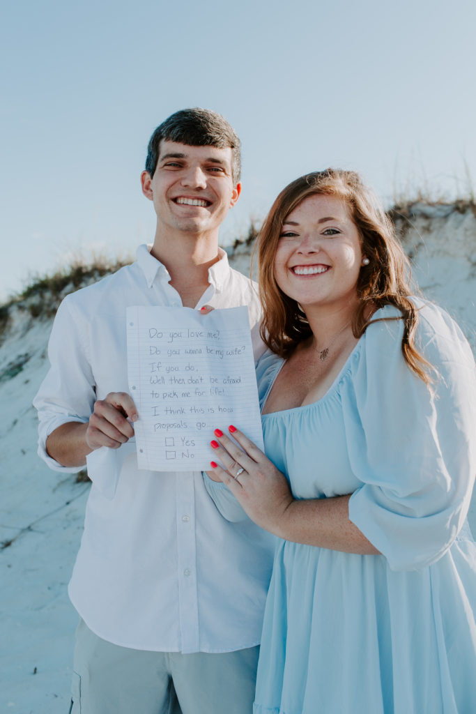 Woman and man smiling as they are holding up the note he proposed with on a beach in Destin
