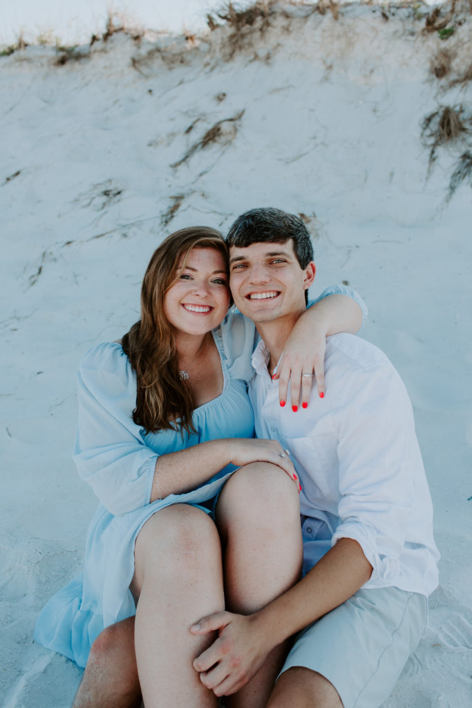 Woman hugging on her partner with her arm around his neck as they are sitting on the beach during their beach engagement