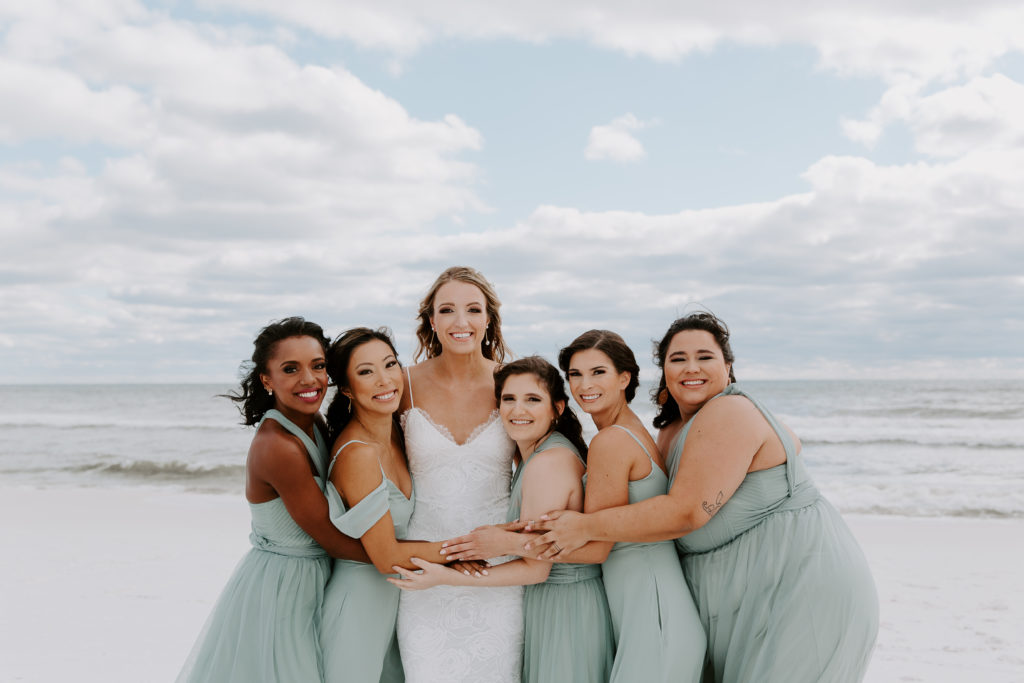Bridesmaids hugging on the bride as they are all standing on the beach smiling during a Destin wedding