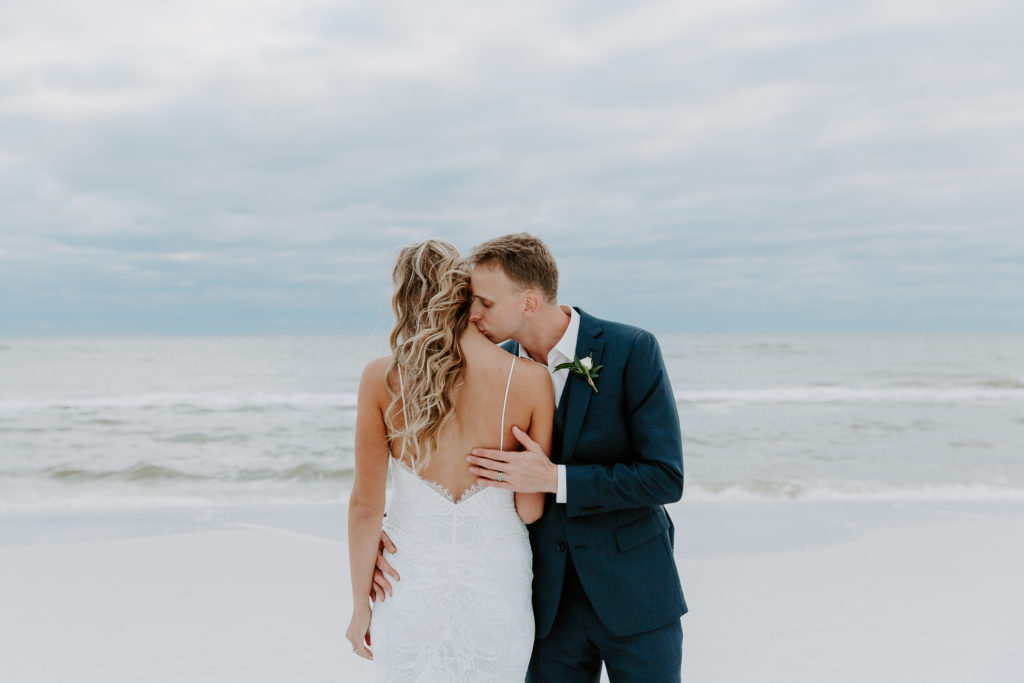 Man kissing his partners neck as she is facing the ocean and the wind is blowing her hair during their Miramar Beach wedding