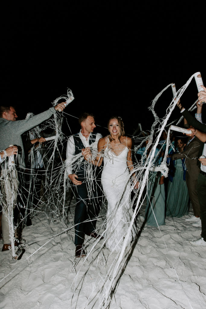 Man looking at his bride as he is holding her hand and they are walking through streamers during their beach exit