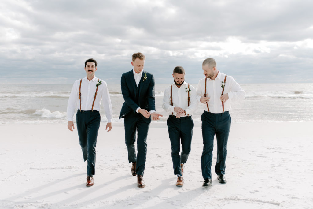 Groom and groomsmen walking away from the ocean and fixing their cufflinks during a Miramar Beach wedding