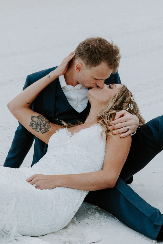 Woman bringing her partner in for a kiss as she is leaning up against him and they are both sitting in the sad during their 30A wedding