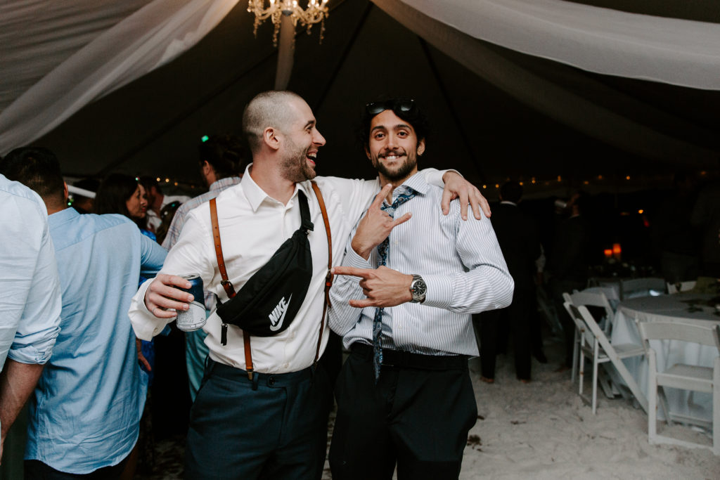 Man throwing his arm around the shoulders of his friend who is throwing up the rocker symbol during a beach wedding reception