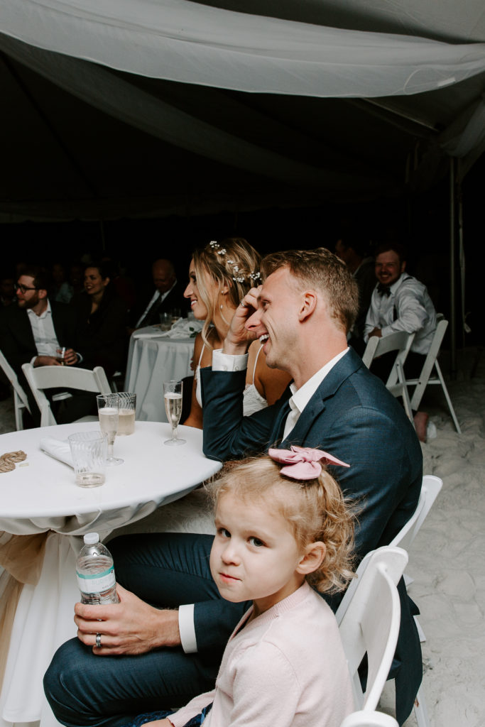 Groom laughing during speeches as his niece is sitting next to him during their beach wedding