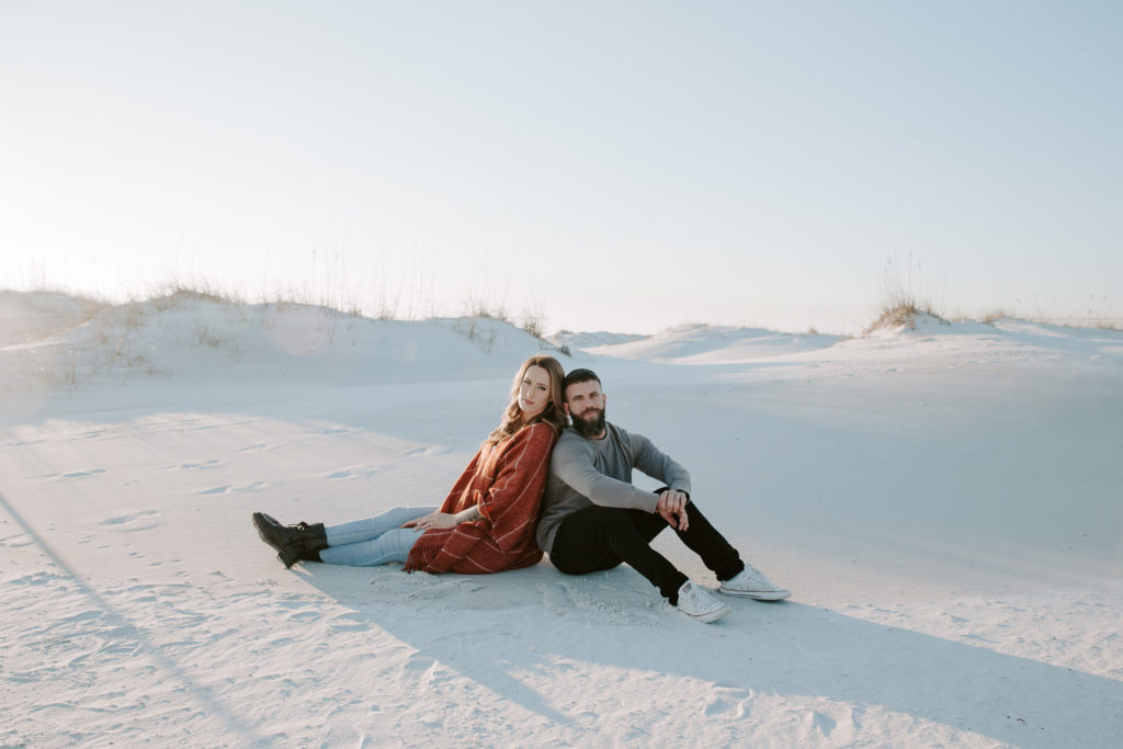 Couple sitting in front of sand dunes as they lean their backs together during their couple photos in Destin
