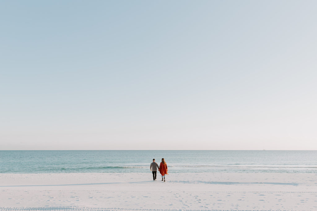 Newly engaged couple holding hands and walking towards the gulf of mexico as the sun is setting