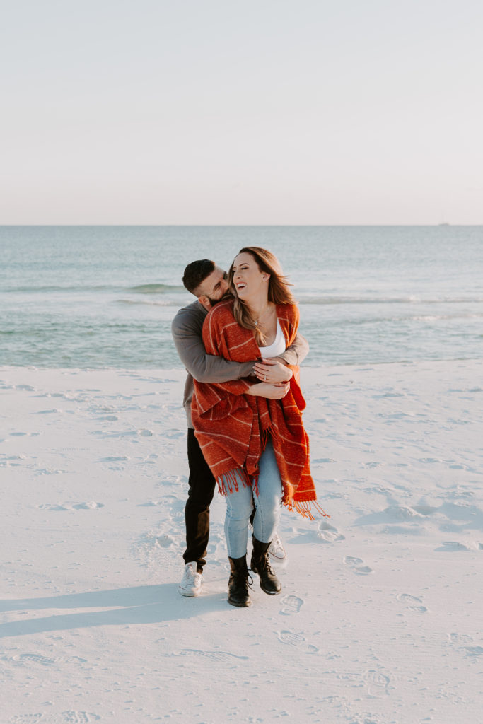 Man holding his partner from the back as she is laughing after he surprised her during their winter beach engagement photos