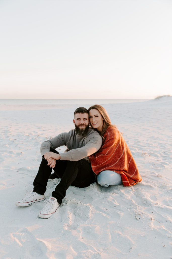 Couple sitting in the sand while the man has his arms resting on his knees and the woman is hugging onto him and they are both smiling with the sun setting during their beach engagement photos