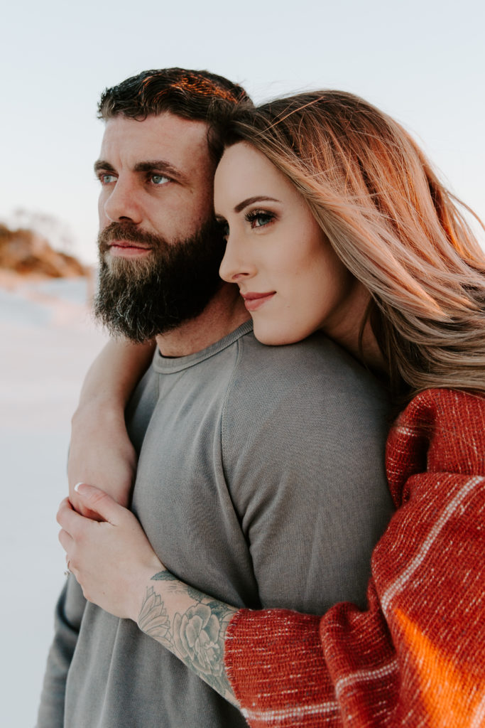 Woman hugging on her partner from behind as she is resting her chin on his shoulder and they are looking off in to the distance during their emerald coast engagement photos