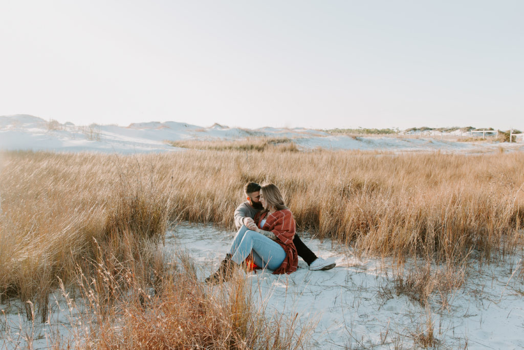 Man and woman sitting in the midst of beach grass and embracing each other as the sun sets in the back during their beach couple photos in Florida