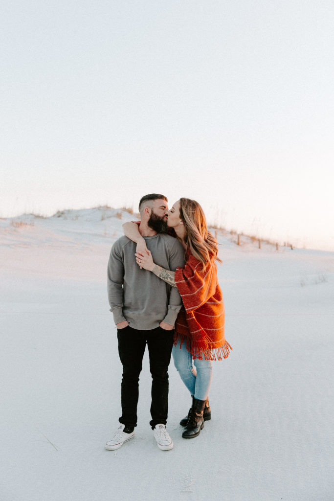 Couple standing in front of sand dunes while the woman has her arms draped around her partner and they are sharing a kiss during their beach sunset engagement photos in Florida