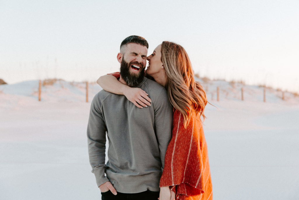 Man laughing as his partner is whispering into his ear and holding his hand with sand dunes in the background during their engagement photos in Florida