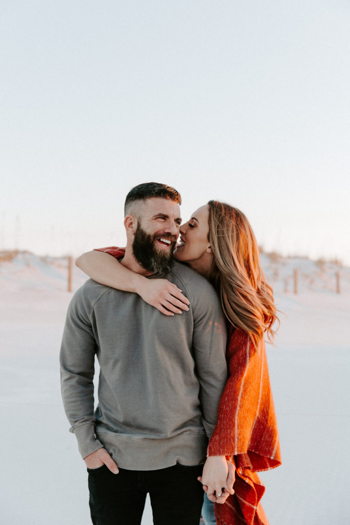 Woman holding onto her partners hand as she whispers into his ear and he is laughing during their beach engagement photos in Florida