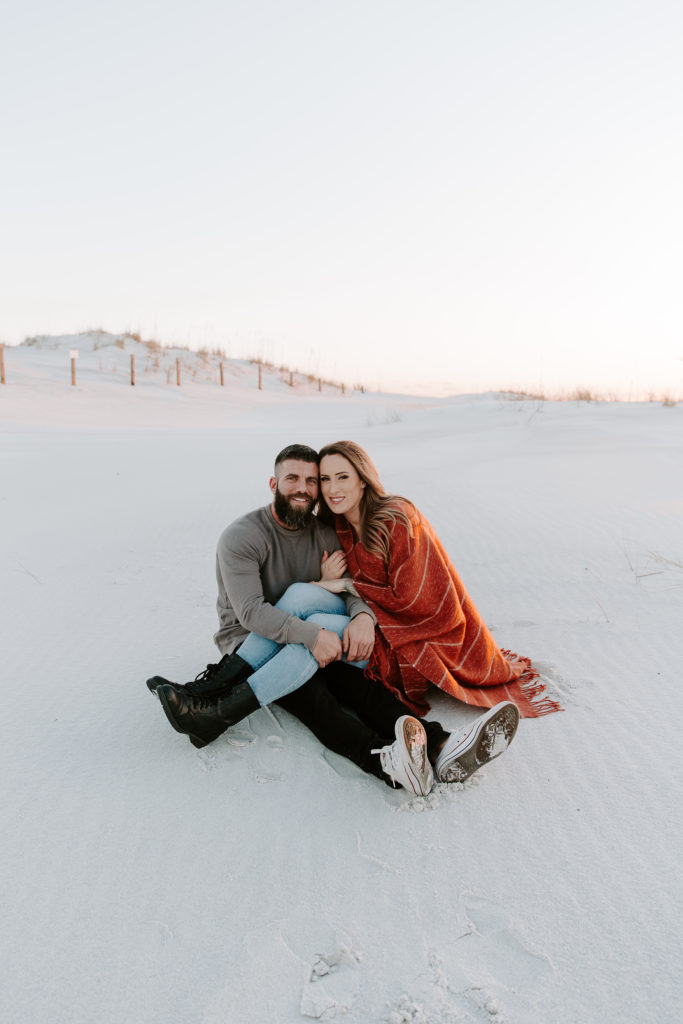 Man and woman sitting in the sand with the woman's legs over the mans as they are smiling and holding on to each other during their Florida engagement photos