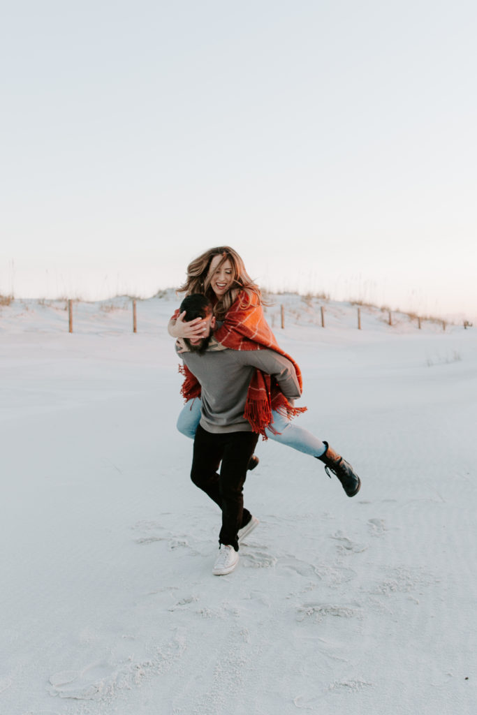 Man giving his partner a piggy back ride as she is blinding him with her hands on accident during their beach engagement photos in the Florida panhandle