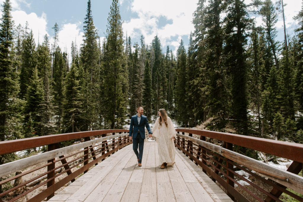 Couple walking along a bridge surrounded by pine trees as they are looking and laughing at each other
