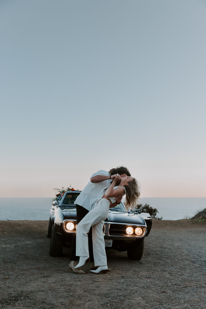 Man dipping his partner back as she is smiling and they are standing in front of a car with its headlights on as dusk settles in during their cliff side engagement photos in California